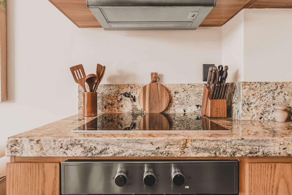 A modern kitchen features a black induction cooktop integrated into a granite countertop, with wooden cabinetry below and a stainless steel range hood above. Wooden utensils and a knife block are neatly arranged on the counter.