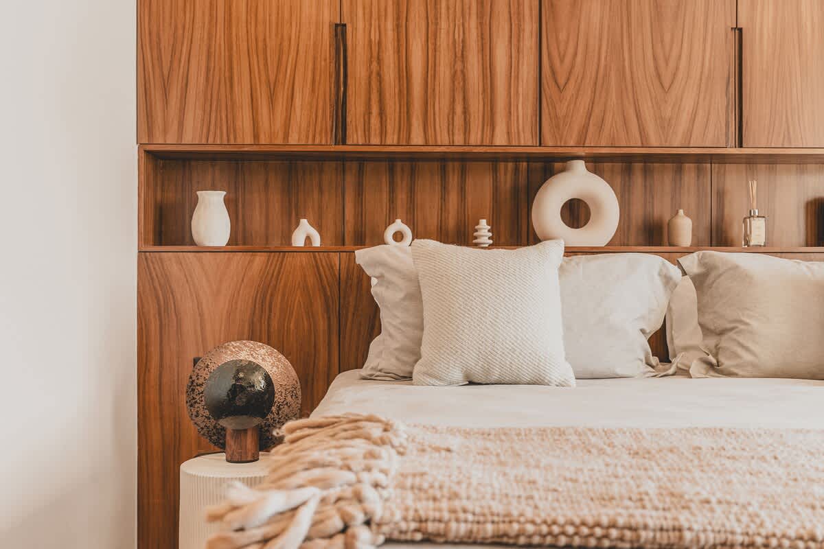 A close-up of the bedroom shows a large bed with neutral-toned bedding and decorative pillows, set against a rich wooden headboard with integrated shelving holding various white ceramic decor. A unique spherical lamp sits on a white fluted side table.