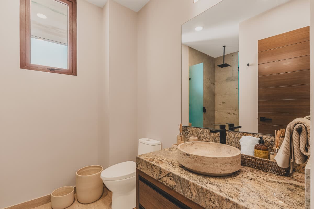 This bathroom features a light-colored stone vessel sink on a granite vanity, with a large mirror and a toilet visible. A small window provides natural light, and wooden accents are present.
