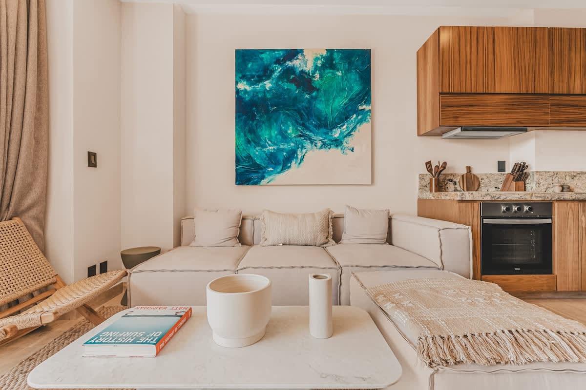 A cozy living room features a light-colored sectional sofa with throw pillows, a white coffee table with decorative vases, and a vibrant abstract painting, with a partial view of the modern kitchen in the background.