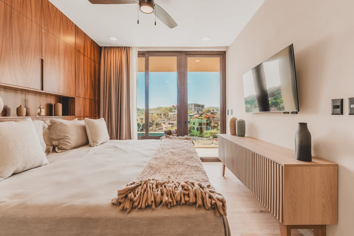 Master bedroom with a large bed, light bedding, and a textured throw, facing a wall-mounted television above a slatted wooden console, with sliding glass doors leading to a balcony.