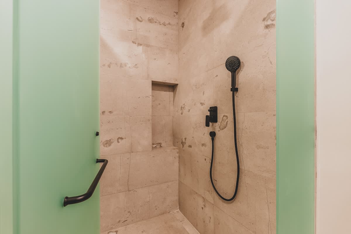 Modern walk-in shower with light-colored stone tiles, a built-in niche for toiletries, and a sleek black rainfall showerhead and handheld sprayer, enclosed by a frosted glass panel.