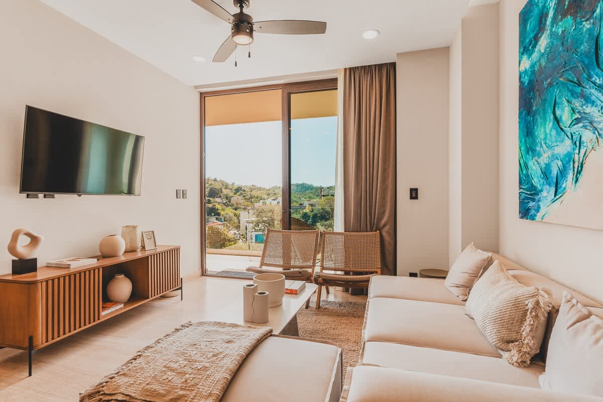 The modern living room features a large wall-mounted TV above a wooden console, a light-colored sectional sofa, and two woven chairs, with sliding glass doors opening to a balcony offering jungle and city views.