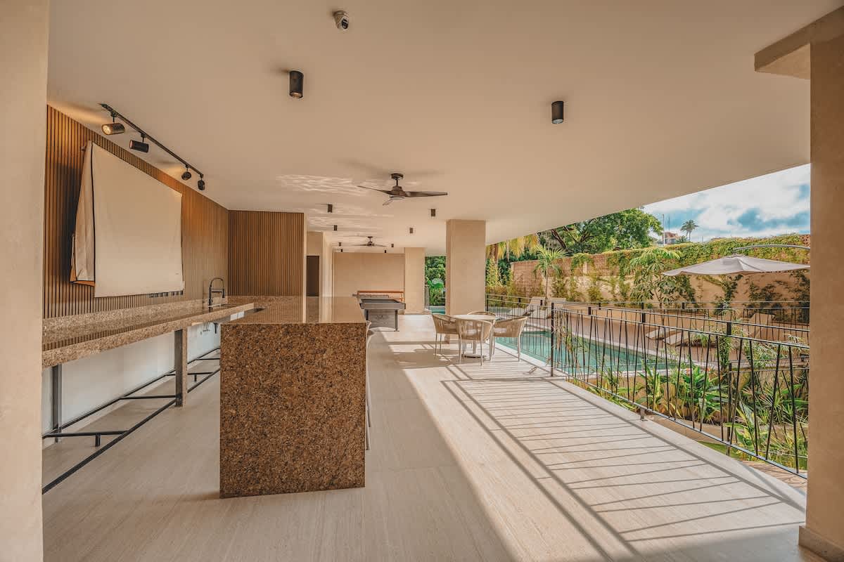 Covered outdoor lounge area next to a swimming pool, featuring a long granite-topped bar with a sink, a large island, and a dining table with chairs. The space has light-colored tile flooring and overlooks lush greenery.