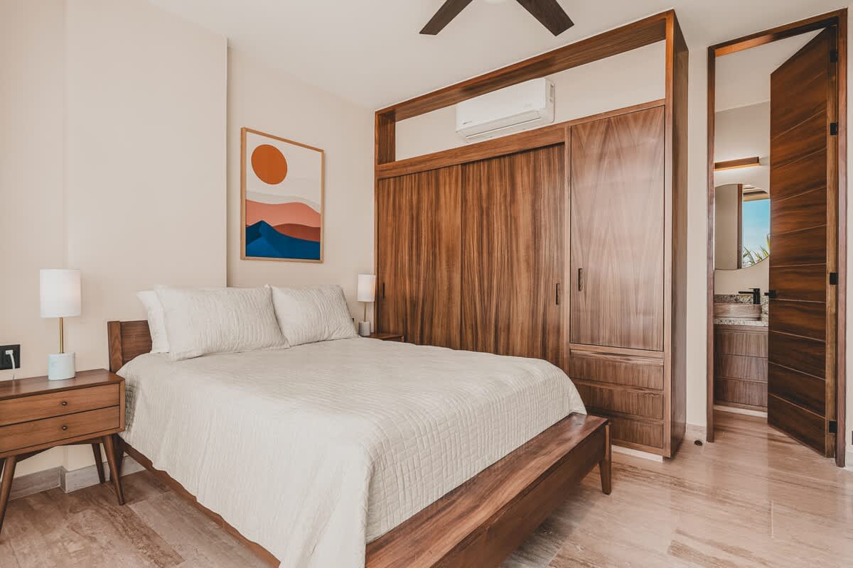 Bright bedroom featuring a queen-sized wooden bed with light bedding, two nightstands, and a large built-in dark wood wardrobe. An abstract artwork adorns the wall, and a doorway leads to an en-suite bathroom.