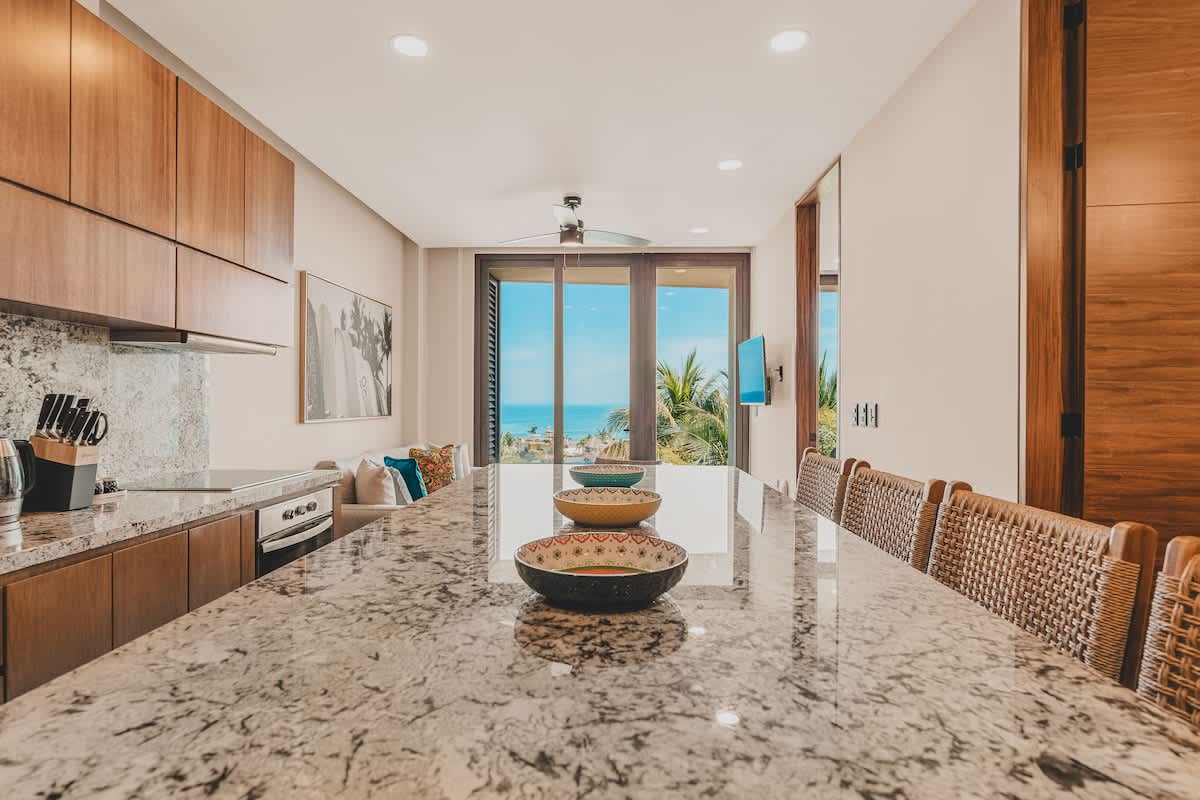 Modern kitchen featuring a long granite-topped island with integrated seating, light wood cabinetry, and a clear sightline through the living area to the ocean beyond, creating an open and airy feel.