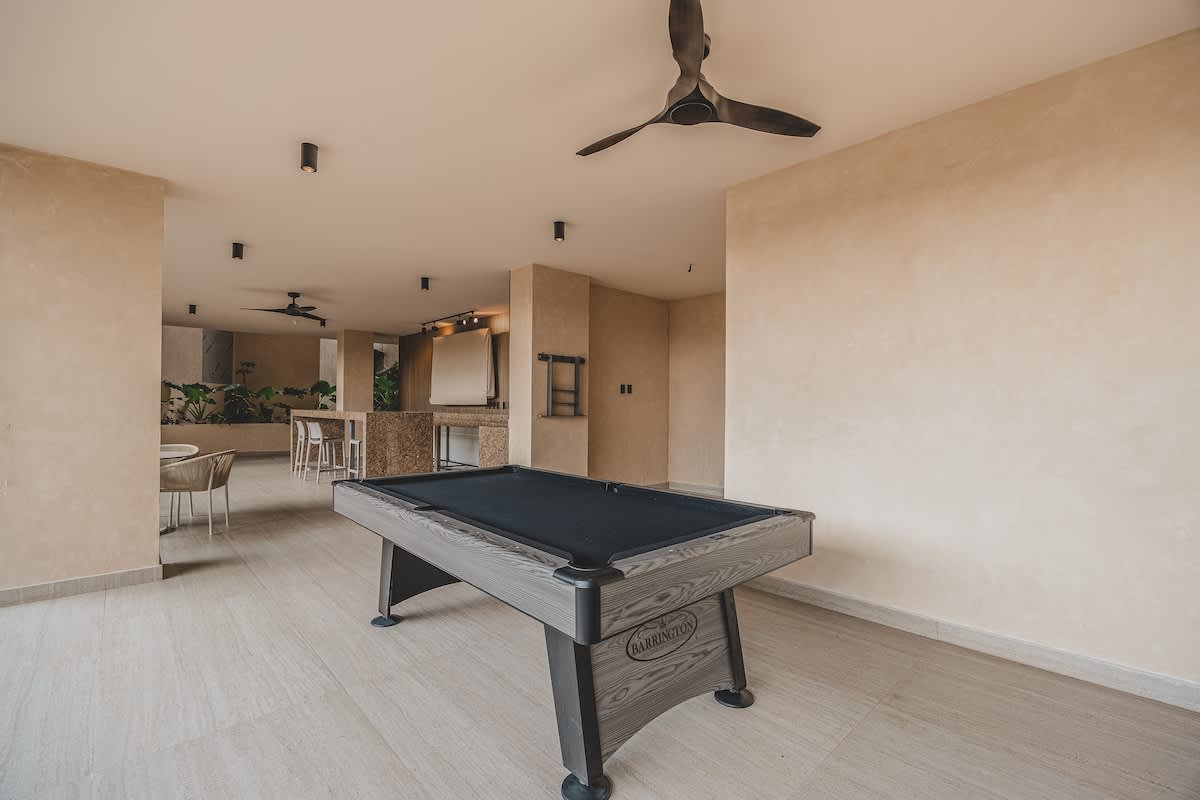 A black-felted pool table with a grey wood frame is centrally located in a spacious covered outdoor game area with light tiled flooring. A ceiling fan and recessed lighting illuminate the space, with light-colored walls.