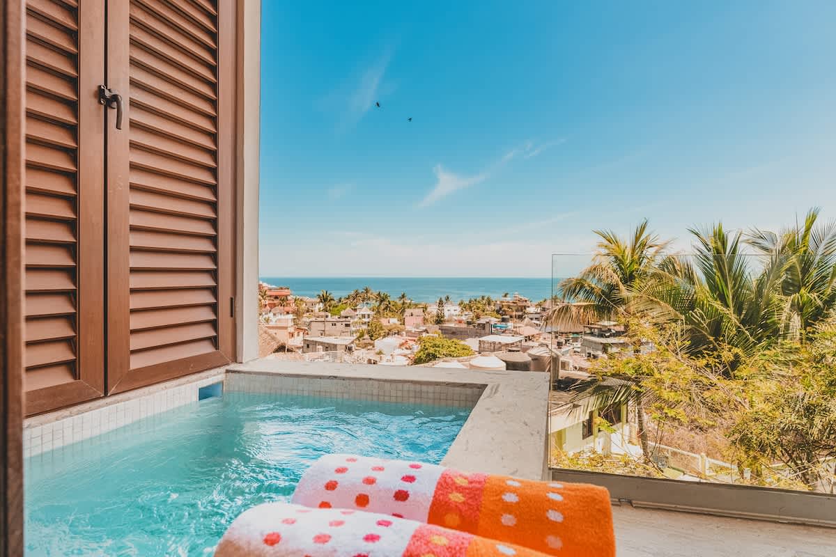 Close-up view of a private balcony hot tub with vibrant blue water, offering a picturesque backdrop of the ocean and a lush tropical town. Two colorful towels are placed on the edge.