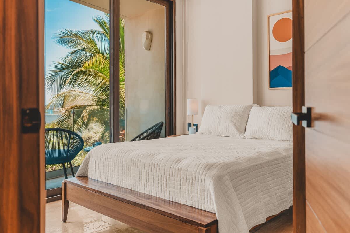 Bright bedroom with a wooden platform bed, white bedding, and a colorful abstract art piece above. Large sliding glass doors open to a private balcony with ocean views and lush palm trees.