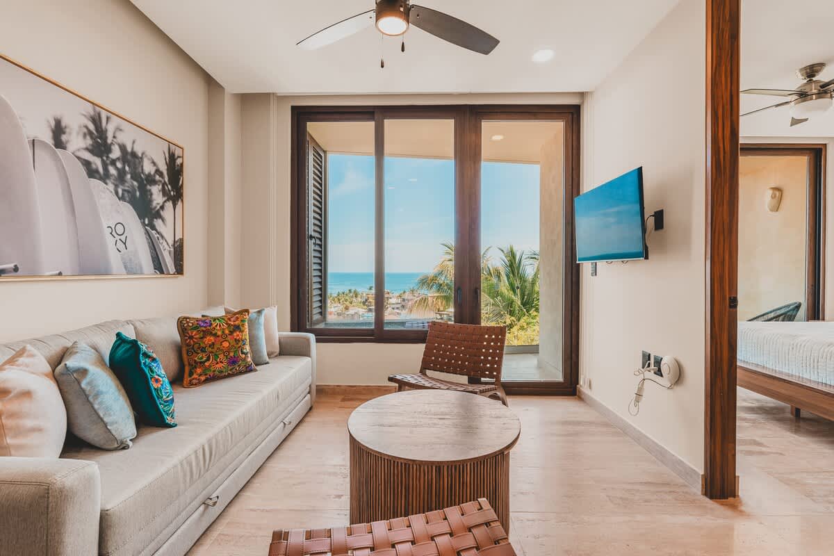 Bright living room with a light grey sofa, two woven leather chairs, and a round wooden coffee table, featuring large windows that open to a balcony with ocean and palm tree views. A ceiling fan and wall-mounted TV are present.