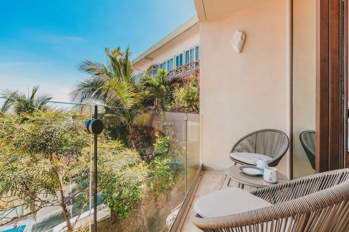 Private balcony with glass railings, furnished with two woven armchairs and a small round table, overlooking lush tropical foliage and distant buildings under a bright blue sky.