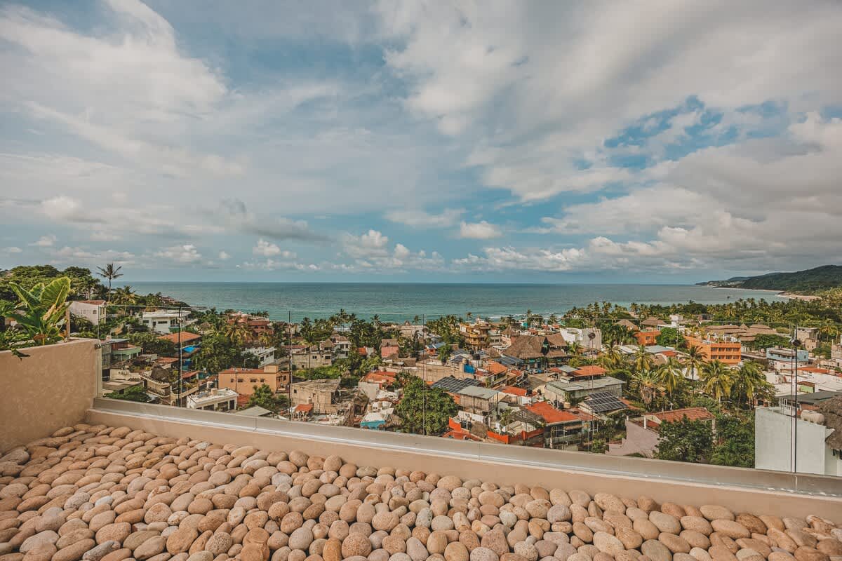 Wide shot of the rooftop deck showcasing the extensive use of light-colored river rocks and a clear glass railing, providing an unobstructed panoramic view of the ocean and the bustling coastal town below. The sky is partly cloudy.