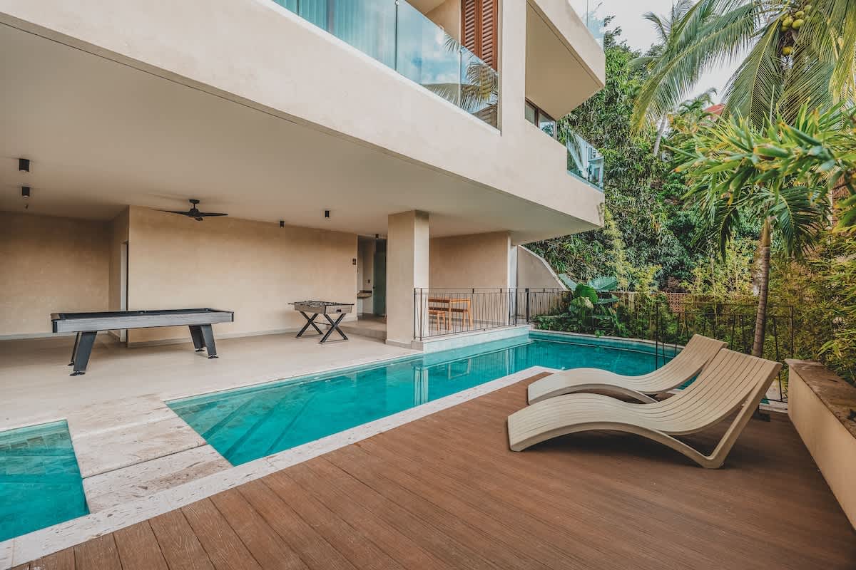 An expansive outdoor recreation area features a turquoise swimming pool bordered by a wooden deck with two lounge chairs. Adjacent is a covered patio housing a pool table and a foosball table, all surrounded by lush tropical greenery and modern architecture.