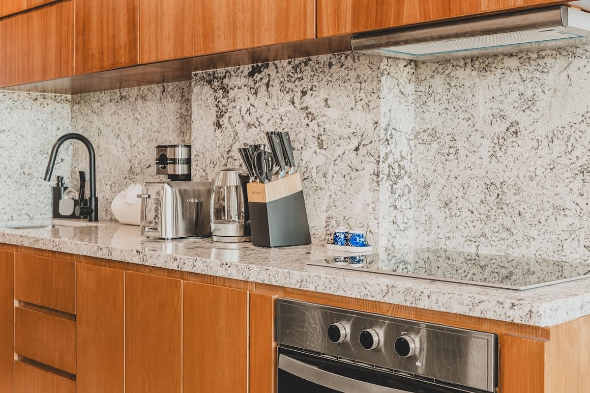 Close-up of a modern kitchen counter with light granite, featuring a built-in electric cooktop, stainless steel oven, and various small appliances. The backsplash matches the countertop, and rich wooden cabinets provide ample storage.