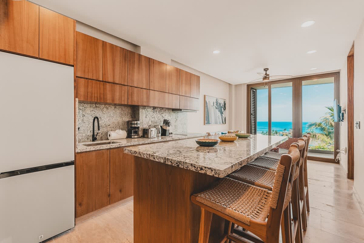 Spacious and modern kitchen featuring a long granite island that doubles as a dining bar with woven bar stools, ample light wood cabinetry, and an open layout connecting to the living area with ocean views.