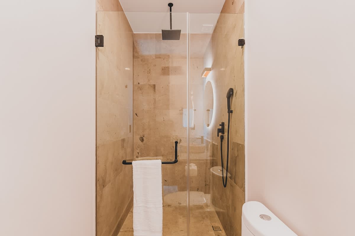 Contemporary bathroom featuring a spacious glass-enclosed shower with a rainfall showerhead and handheld sprayer, set against light stone-tiled walls, next to a white toilet.