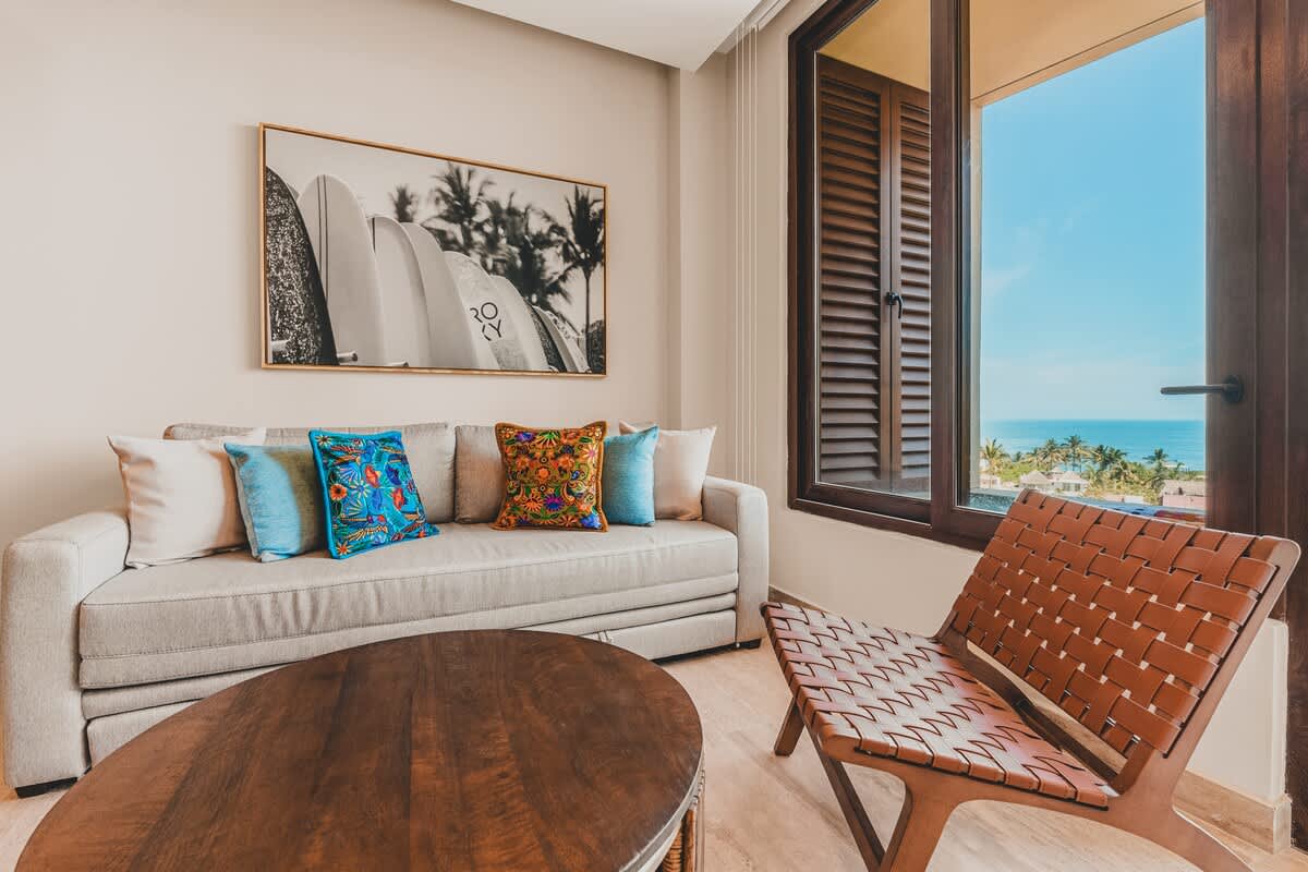 Inviting living room with a light-colored sofa adorned with colorful throw pillows, a round wooden coffee table, and a woven leather chair. A large window offers expansive ocean views.