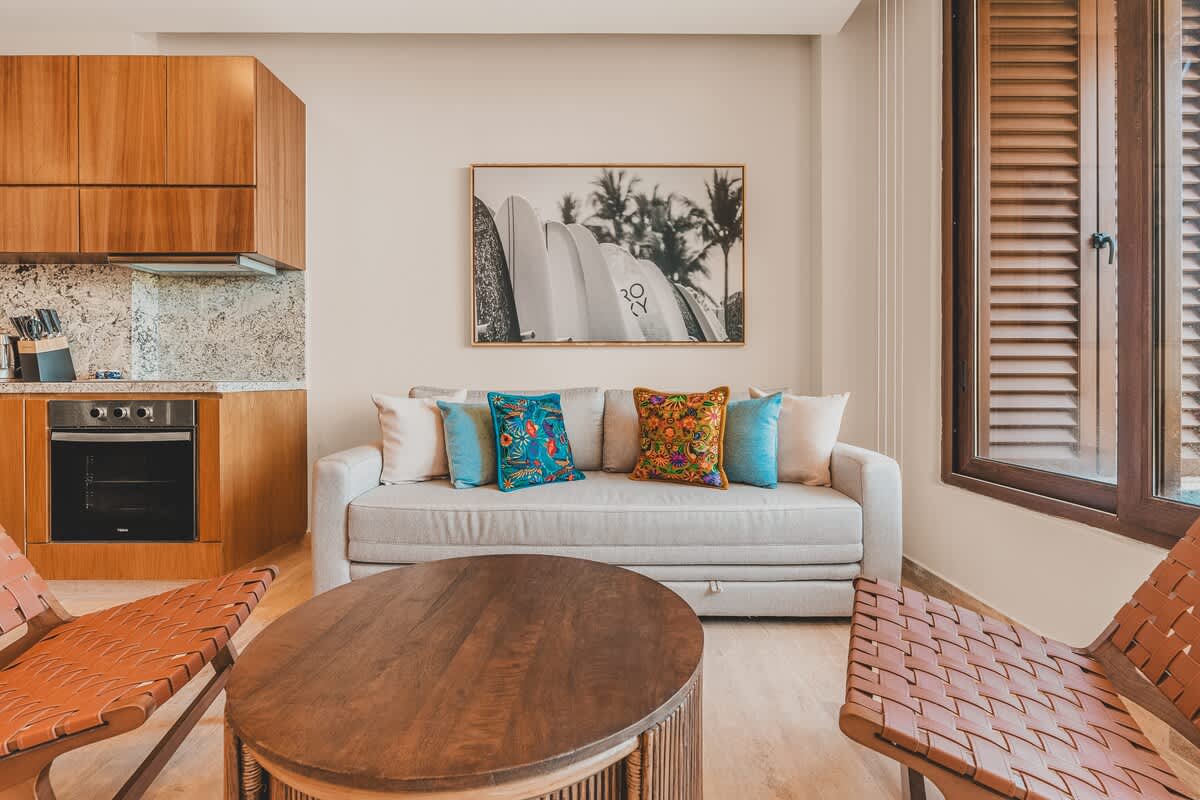 Open-concept living area with a light grey sofa and colorful pillows facing a round wooden coffee table, adjacent to a modern kitchen with wooden cabinetry and granite countertops. A large surfboard art piece adorns the wall.
