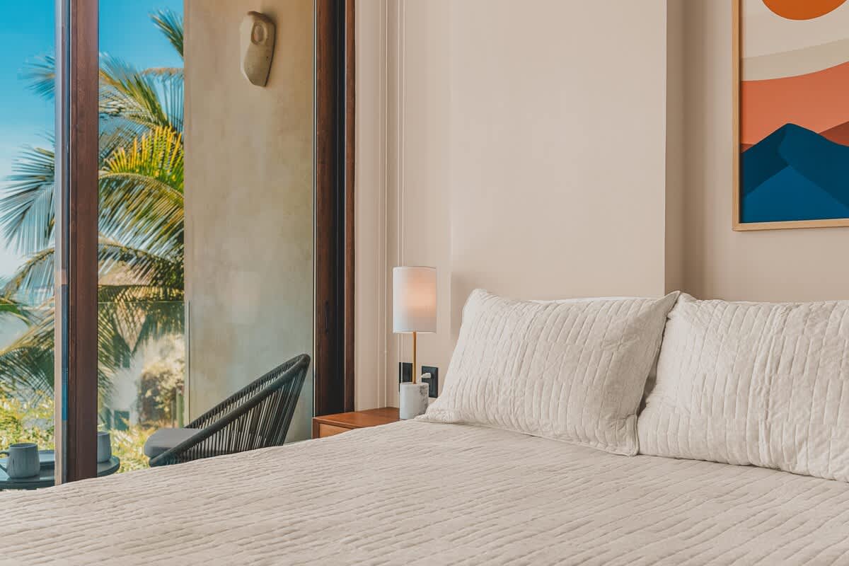 Cozy bedroom with a light-colored quilted bedspread and pillows, featuring a wooden headboard and nightstand with a lamp. A large glass door opens to a private balcony with tropical views.