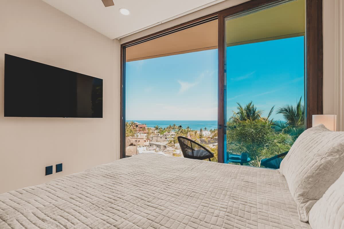 Bright bedroom featuring a neatly made bed, a wall-mounted flat-screen TV, and large sliding glass doors that open to a balcony with expansive ocean and town views.