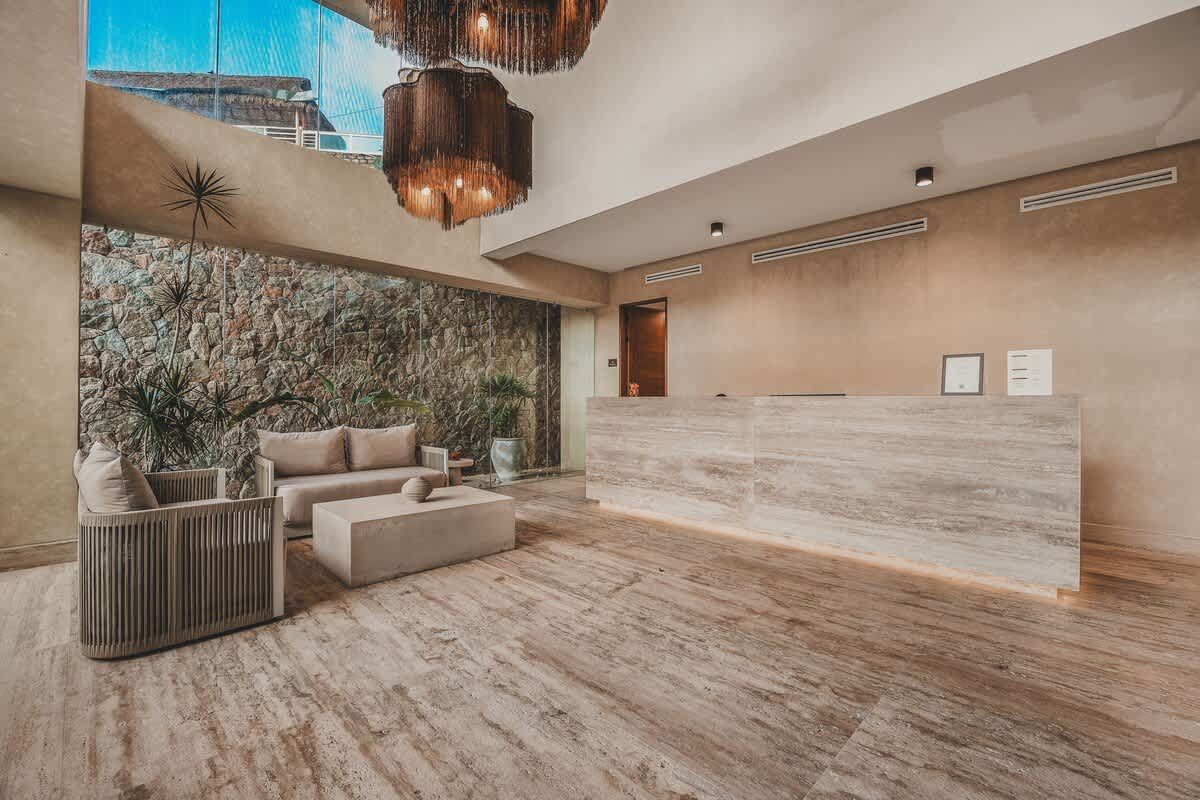 A welcoming lobby space featuring a long, light-toned reception desk with subtle underlighting, and a comfortable seating area with a sofa and armchair. The room is characterized by a textured stone wall, high ceilings, and elegant decorative chandeliers.