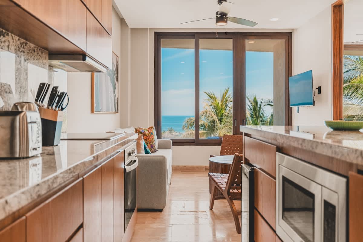 Bright and airy living space with a comfortable sofa and a small dining table, offering stunning ocean views through large sliding glass doors. The kitchen counter with a microwave is visible on the right.