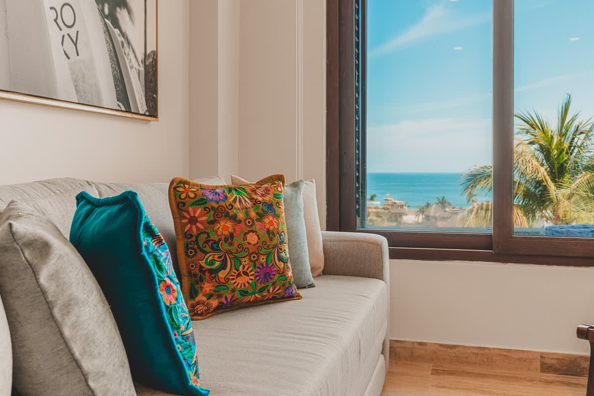 Cozy living room featuring a light gray sofa adorned with colorful, embroidered pillows, positioned next to a large window offering expansive ocean and palm tree views.