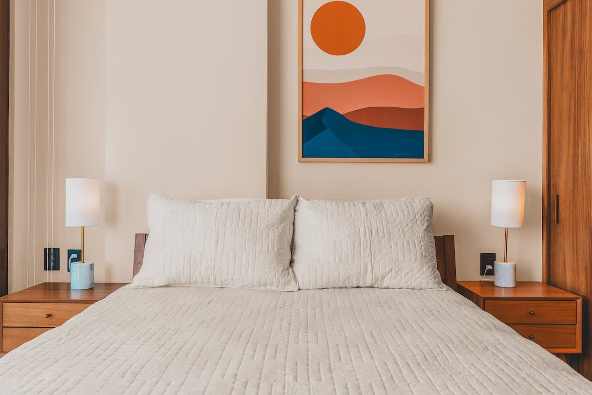 Symmetrical view of a bedroom with a queen-sized bed, light quilted bedding, and two wooden nightstands each with a modern lamp. A colorful abstract art piece hangs above the bed.