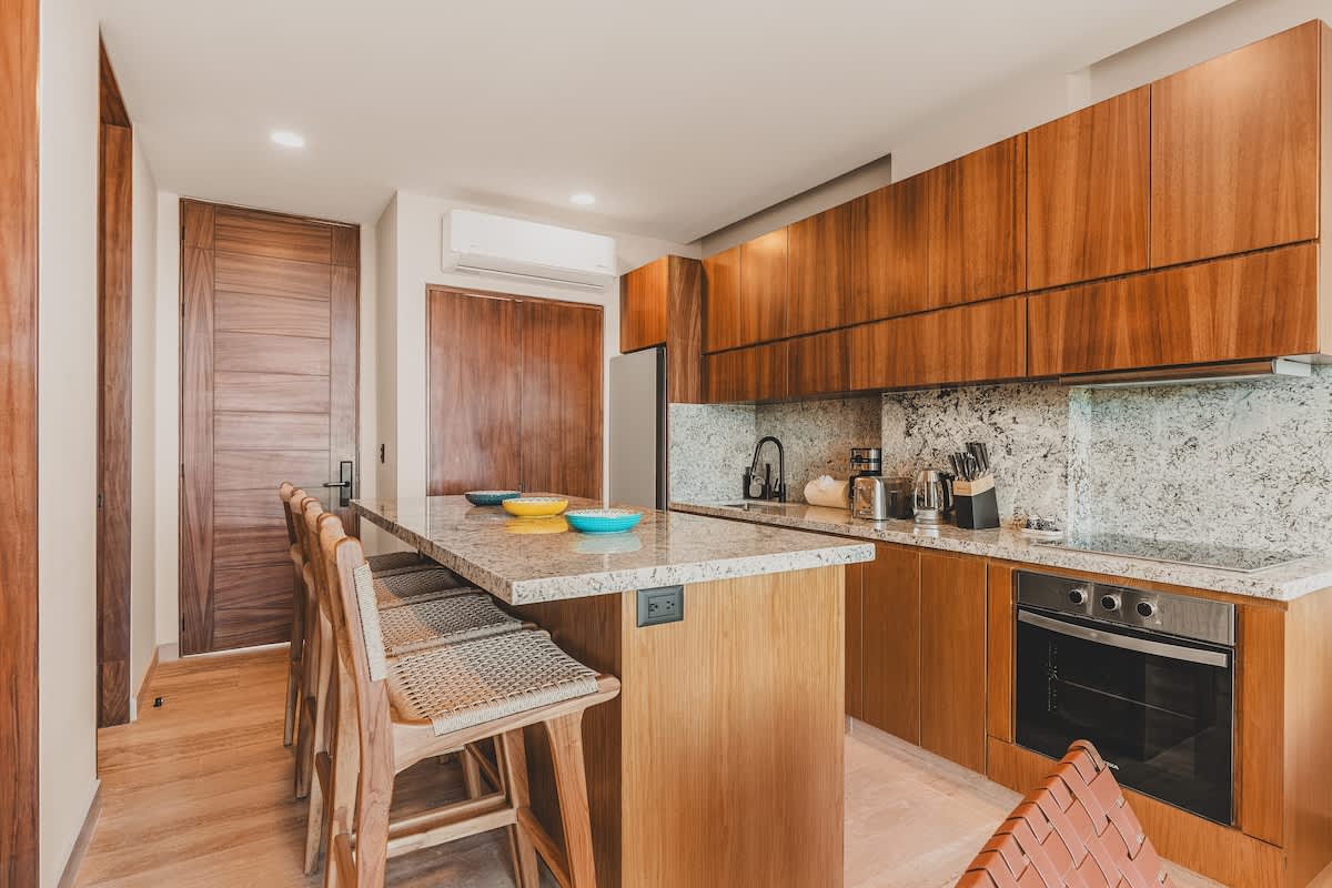 Modern kitchen with rich wooden cabinetry, light granite countertops and backsplash, and a large island with bar seating for four. Stainless steel appliances and a black gooseneck faucet add contemporary touches.
