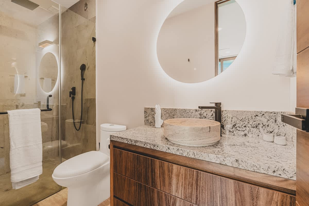 Modern bathroom featuring a glass-enclosed shower with a dark fixture, a white toilet, and a vanity with a light-colored stone countertop and a round vessel sink. A large round backlit mirror hangs above the sink.