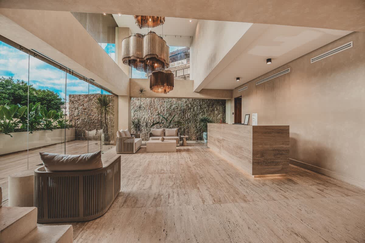 A bright and airy lobby with a long, light-colored reception desk and a seating area with a sofa and armchair. The space features high ceilings, large glass windows, and a stone accent wall, creating an inviting and modern aesthetic.