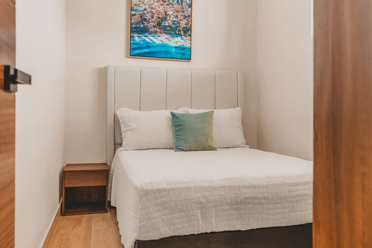 Cozy bedroom featuring a queen-sized bed with a light grey upholstered headboard and white bedding, complemented by a wooden nightstand and a vibrant framed artwork above the bed.