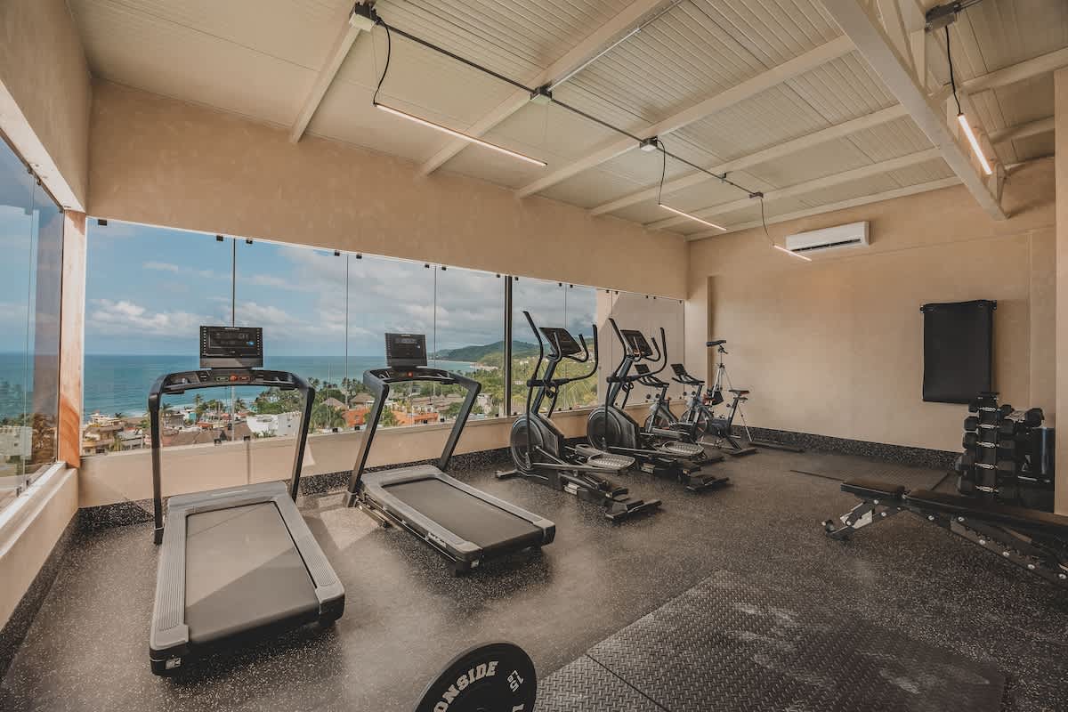 Modern gym with large windows offering panoramic ocean and city views, equipped with two treadmills, an elliptical, and a stationary bike. The space features a black rubber floor and exposed ceiling beams.