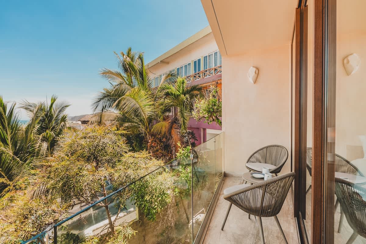Spacious balcony with modern grey woven chairs and a small table, surrounded by lush tropical foliage and offering a glimpse of the building's architecture.