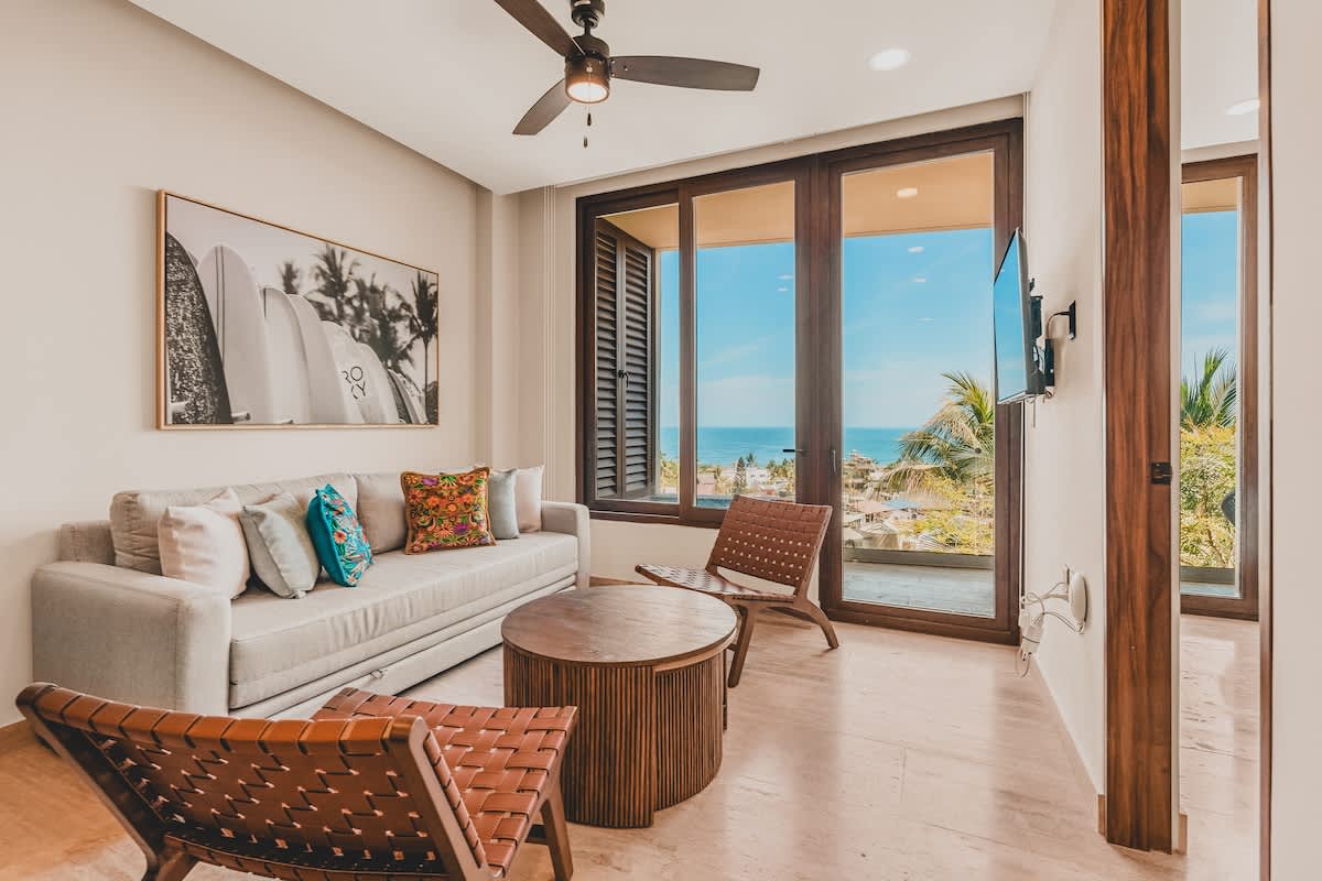 Modern living room featuring a light grey sofa with colorful pillows, two brown woven leather chairs, and a round wooden coffee table, with large windows revealing ocean views. A large framed surfboard art piece hangs on the wall.