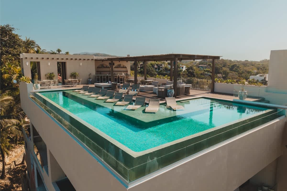 Expansive rooftop area featuring a large infinity pool with submerged lounge chairs, surrounded by comfortable outdoor seating and dining areas under a pergola. The space offers sweeping views of the lush landscape and ocean.