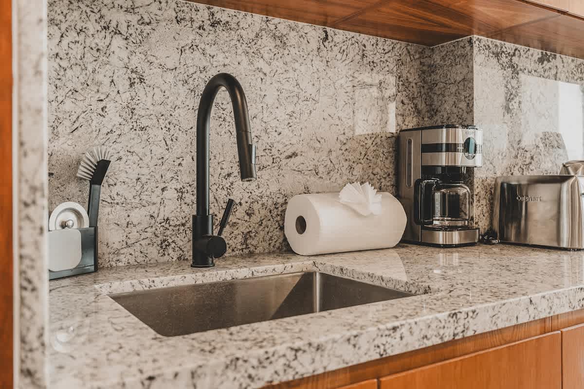 Detailed view of the kitchen sink area, showcasing a deep stainless steel sink with a modern black gooseneck faucet set against a light granite countertop and backsplash. A coffee maker and paper towel roll are visible on the counter.