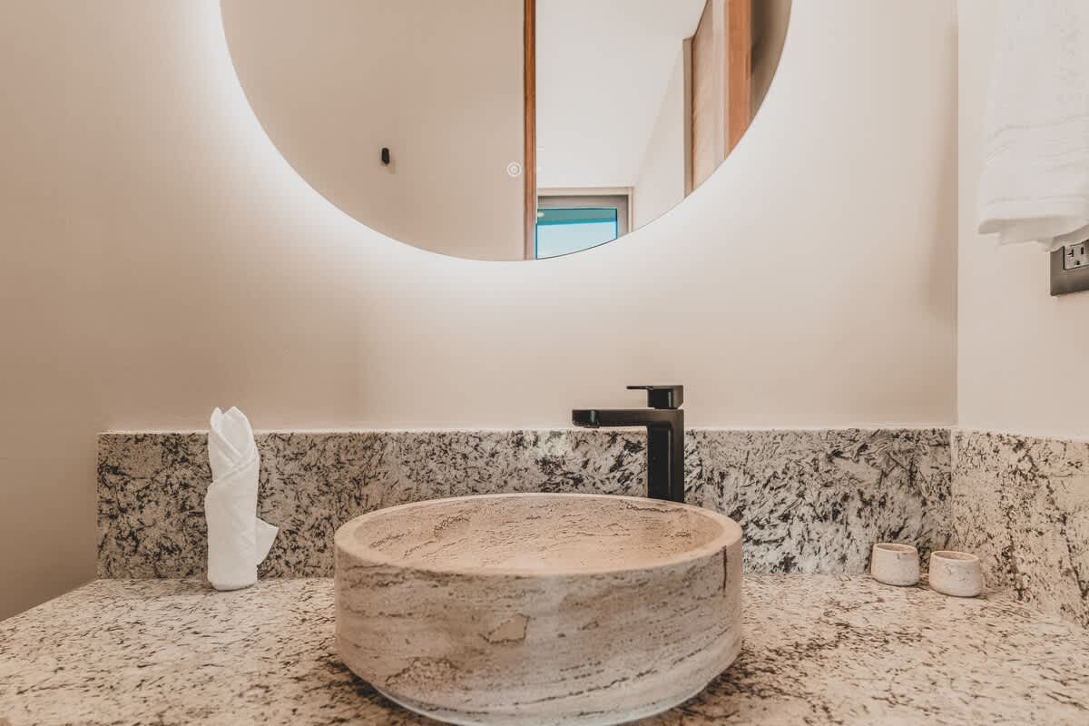 Close-up of a contemporary bathroom vanity with a light-colored, textured stone vessel sink and a black modern faucet, set on a speckled granite countertop. A large round backlit mirror illuminates the space.