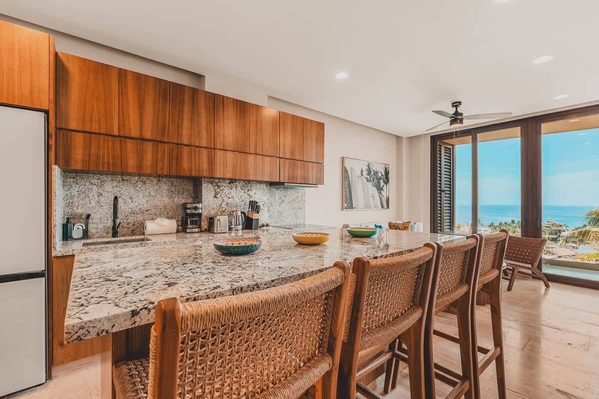 Spacious open-concept kitchen with a large granite island featuring woven bar stools, dark wood cabinetry, and a backsplash matching the countertop. Floor-to-ceiling glass doors offer ocean views.
