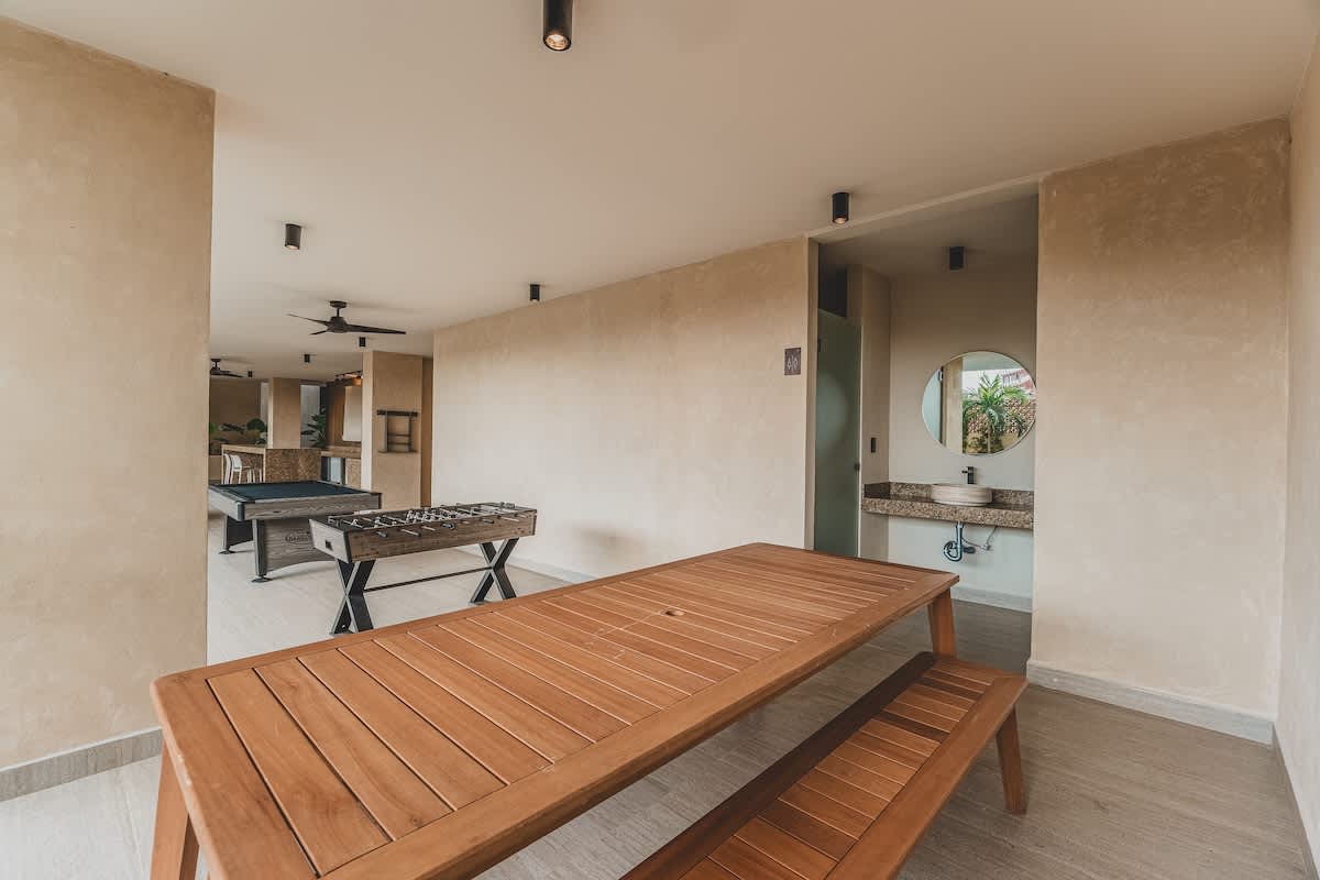 A covered outdoor recreation space features a long wooden picnic table with benches, a foosball table, and a pool table in the distance. A vanity with a round mirror and sink is visible in an adjacent alcove, all set against light-colored walls.