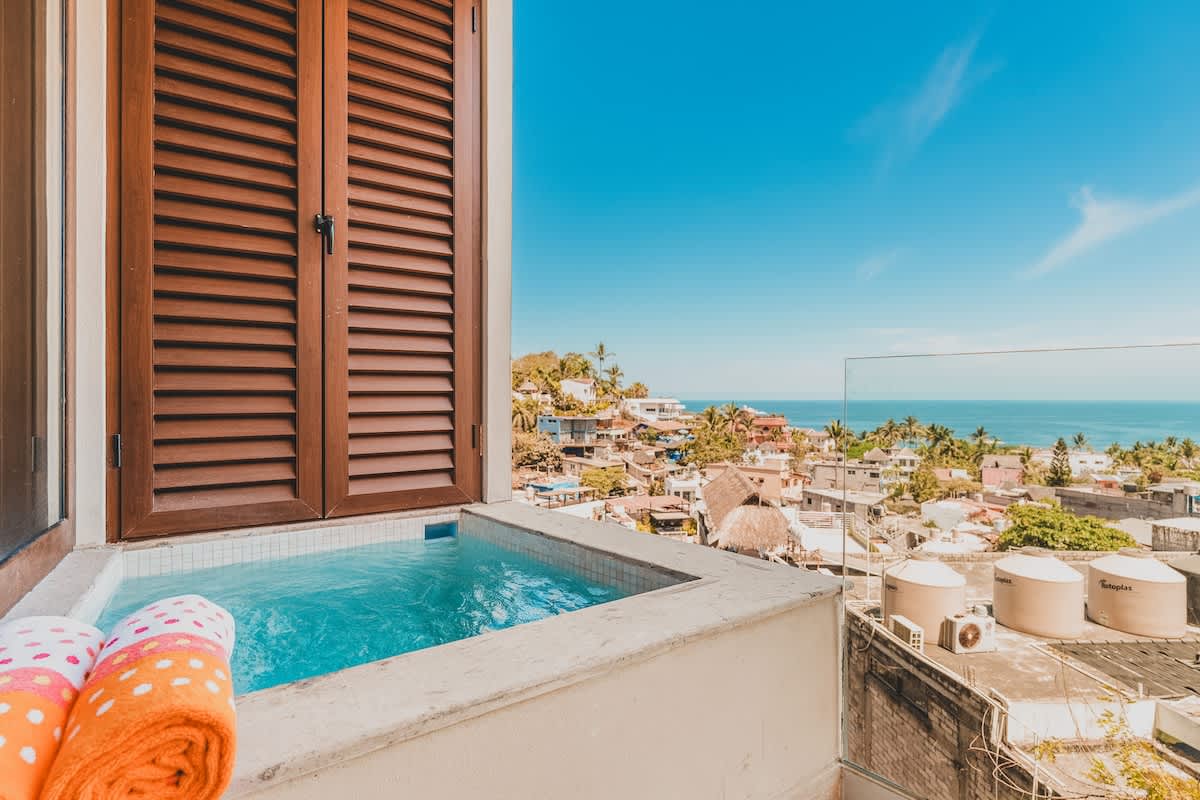 Private balcony hot tub with sparkling blue water, offering expansive views of the town and ocean under a bright blue sky. Two colorful towels are neatly folded beside the tub.