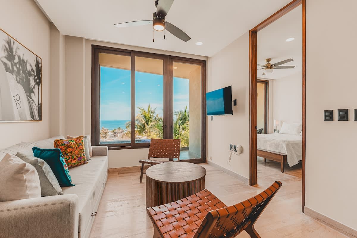 Bright living room with a light grey sofa, two woven leather chairs, a round wooden coffee table, and large windows offering ocean and palm tree views. A ceiling fan and wall-mounted TV are visible.