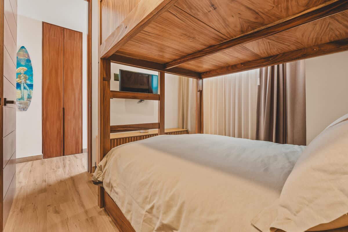 A modern bedroom featuring a wooden bunk bed with a built-in TV shelf, light-colored bedding, and light wood flooring, with a closed wooden door and a decorative surfboard visible in the hallway.