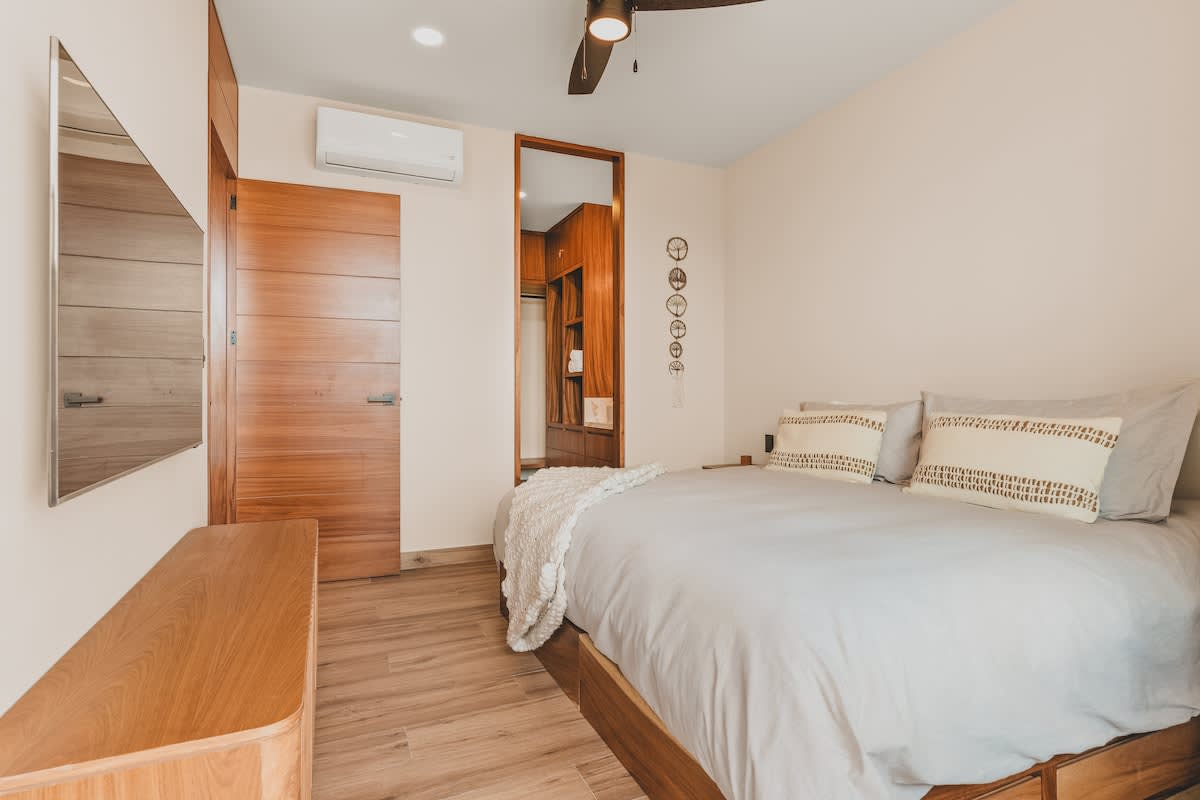 Overview of bedroom-1 showing the queen bed with wooden frame, a wall-mounted TV, an air conditioning unit, and a wooden door leading to a walk-in closet.