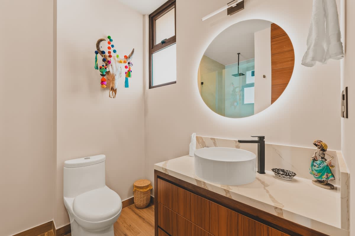 A bright bathroom features a white toilet, a modern vanity with a vessel sink and marble countertop, and a large backlit round mirror, with a colorful decorative skull hanging on the wall.