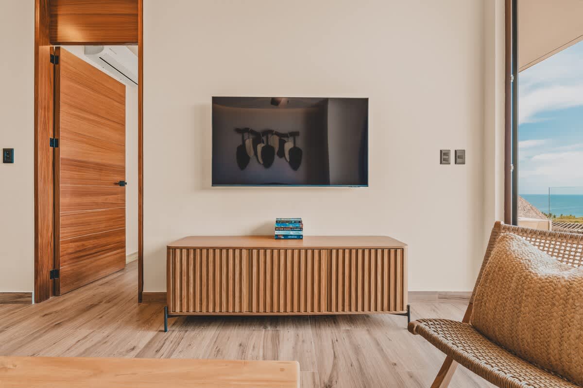 A modern living room featuring a wall-mounted flat-screen TV above a slatted wooden entertainment console. A wooden door is open on the left, revealing another room, and a woven armchair is partially visible on the right.