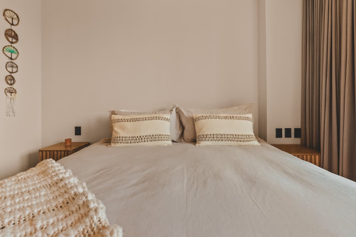 Close-up view of the queen-sized bed in bedroom-1, dressed in light grey linens with two decorative striped pillows, and wooden nightstands on either side.