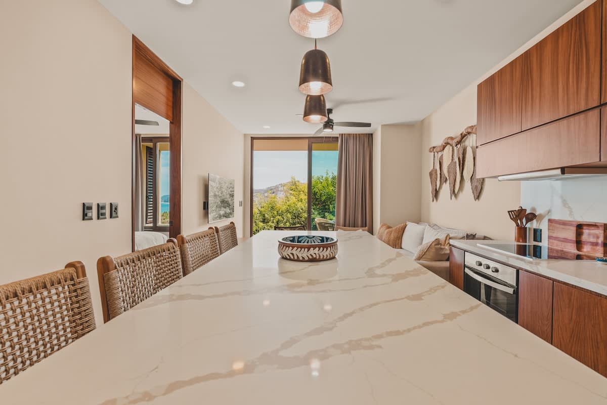 A bright open-concept kitchen and living area with a large white quartz island serving as a dining counter, featuring three woven bar stools. Beyond the island, a living space with a sofa and a balcony with lush greenery and distant hills is visible.