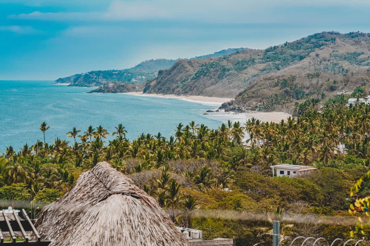 A breathtaking aerial view captures a vast expanse of the Pacific Ocean meeting a sandy beach, framed by lush green hills covered in palm trees and distant coastal homes under a blue sky.