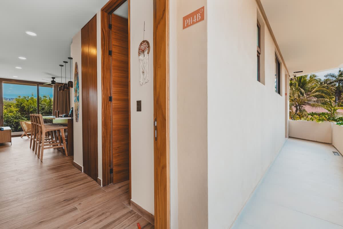 A bright hallway with light wood-look flooring leads past wooden doors and a decorative wall hanging, opening into a living area with large windows and a glimpse of an outdoor view.